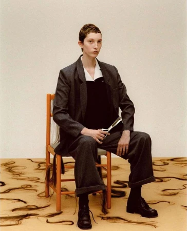 Model with short hair wearing a grey suit and holding scissors seated on an orange chair over scattered hair strands.