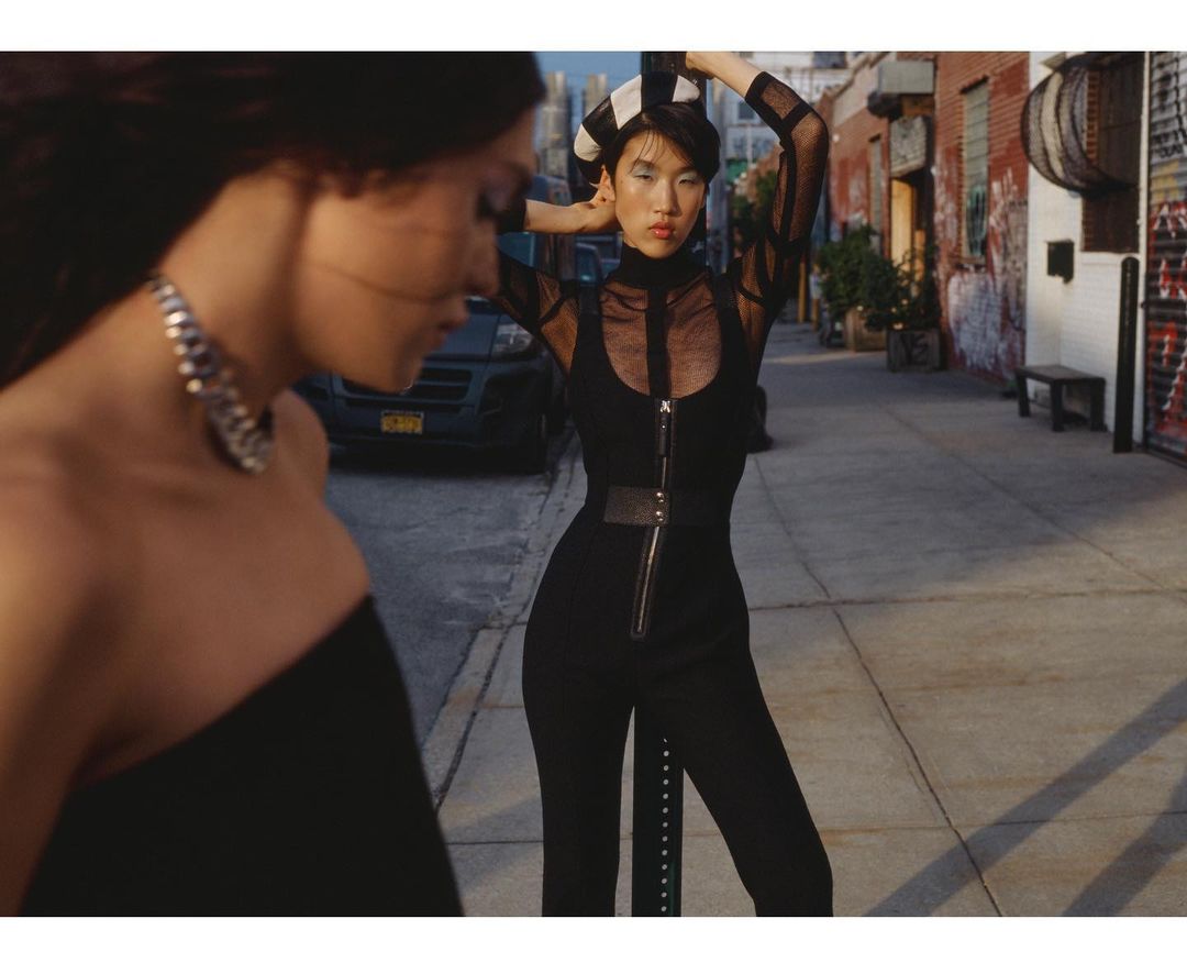 Two women pose on a city street. One woman is out of focus with a silver necklace. The other woman is wearing a black jumpsuit.
