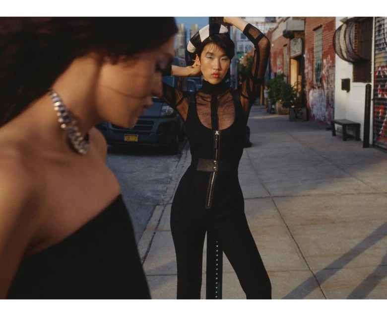 Two women pose on a city street. One woman is out of focus with a silver necklace. The other woman is wearing a black jumpsuit.