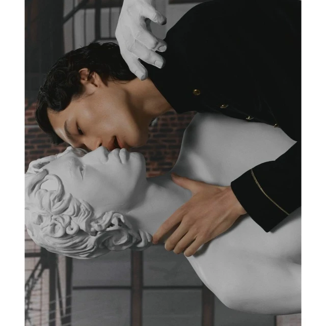 Young adult kisses a white sculpture head, hands with white gloves holding the sculpture. Brick-patterned wallpaper backdrop.