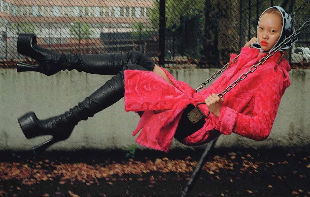 A woman in a pink coat and black boots swings on a playground swing, wearing a headscarf and dark lipstick, set against an urban background.