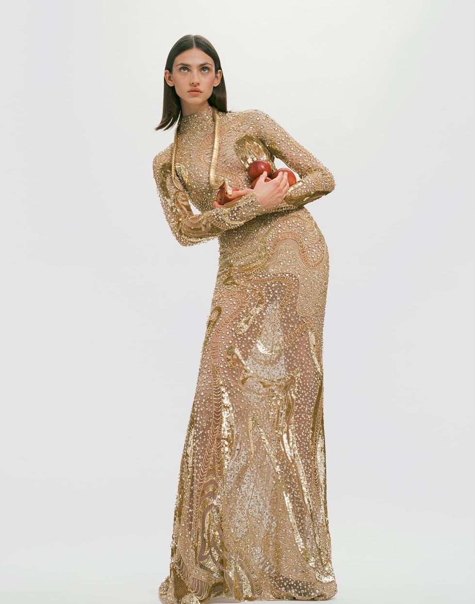 Fashion model in a shimmering gold gown poses with two apples against a plain white backdrop in a studio fashion editorial.