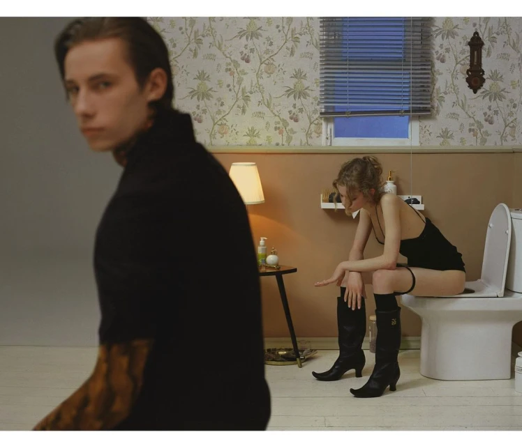 Indoor scene with a man in focus in the foreground and a woman sitting on a toilet in the background, creating a staged and intriguing composition.