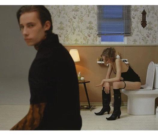 Indoor scene with a man in focus in the foreground and a woman sitting on a toilet in the background, creating a staged and intriguing composition.