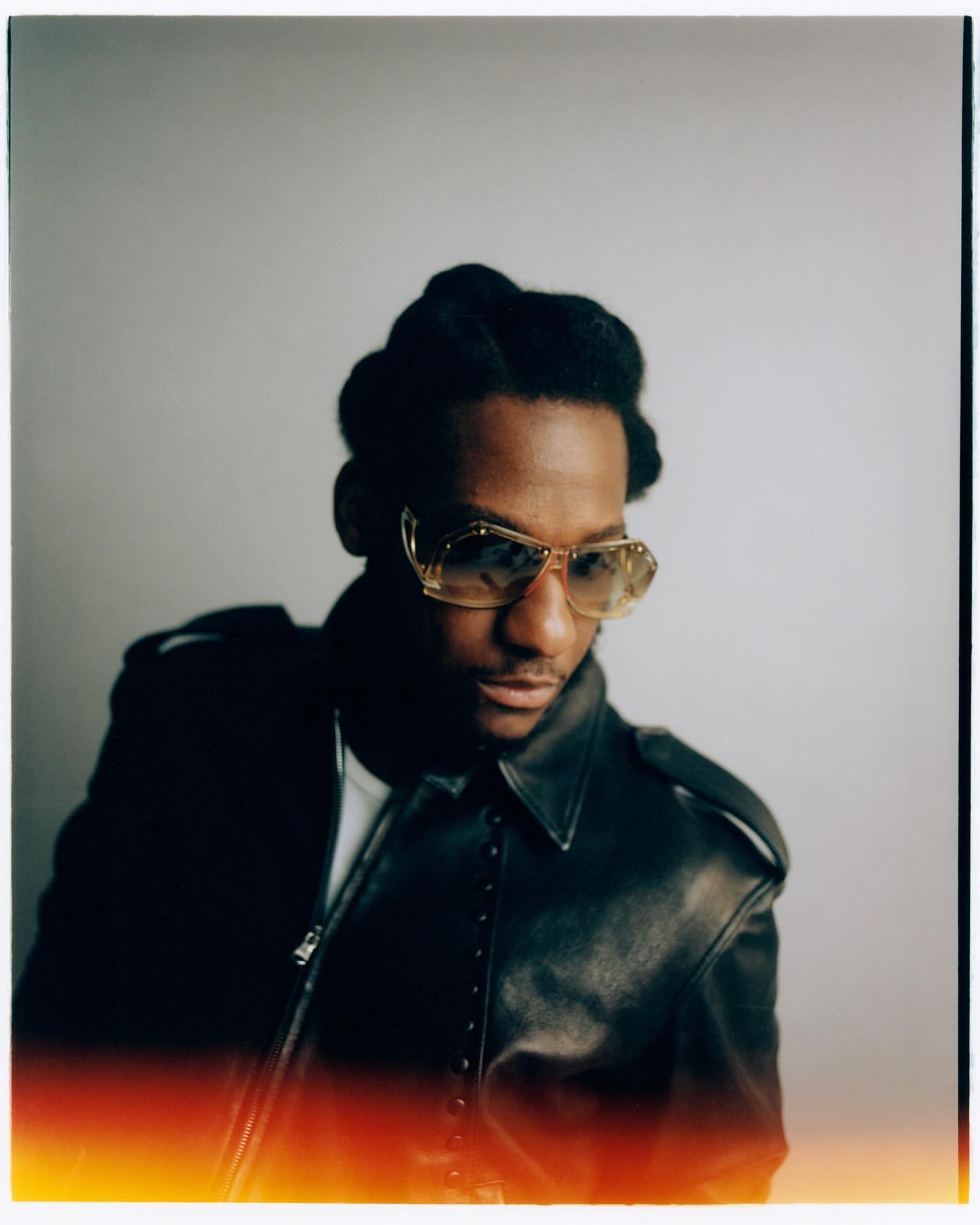 Portrait of Black man with glasses wearing leather jacket against grey background. Studio shot with red vignette along the bottom.