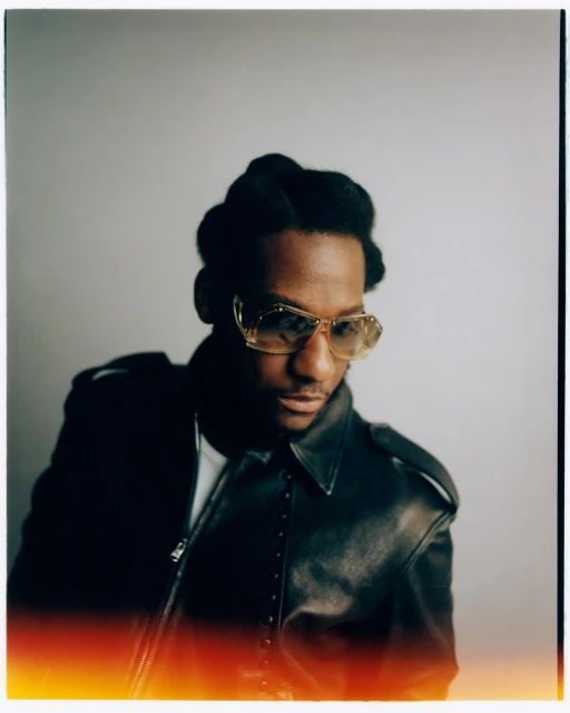 Portrait of Black man with glasses wearing leather jacket against grey background. Studio shot with red vignette along the bottom.