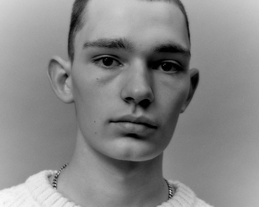Monochrome portrait of a young adult male with dark hair, wearing a knit sweater and chain necklace against a neutral backdrop.