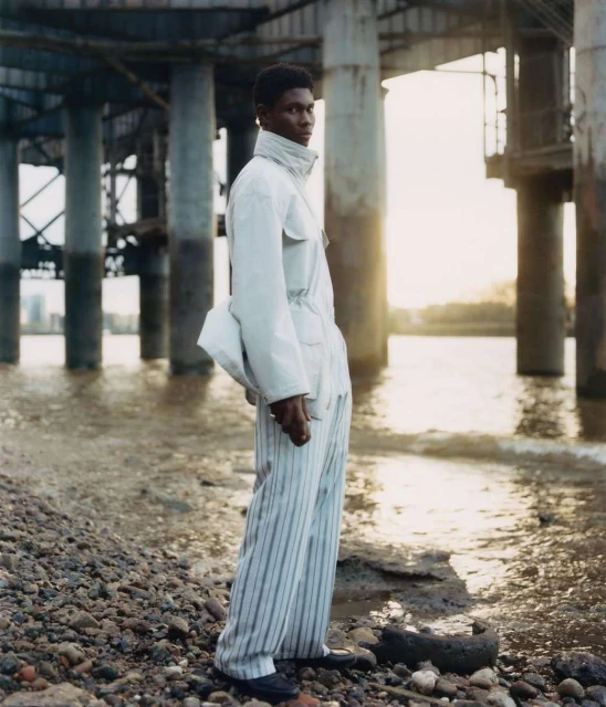 A young black male model poses near water wearing a white jacket and striped pants under concrete pillars for a fashion editorial shoot.
