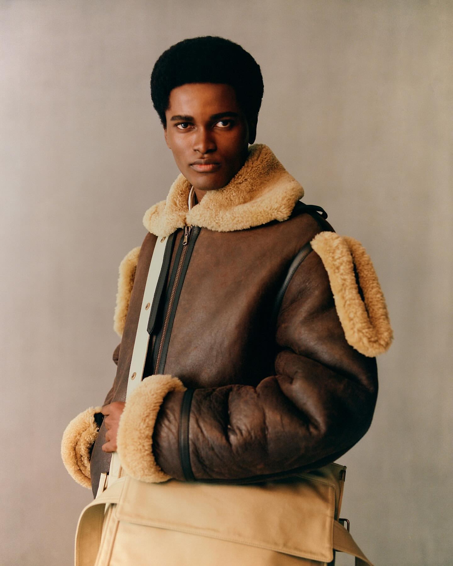 A stylish black male model poses indoors wearing a brown shearling bomber jacket and a beige messenger bag in a fashion editorial shot.