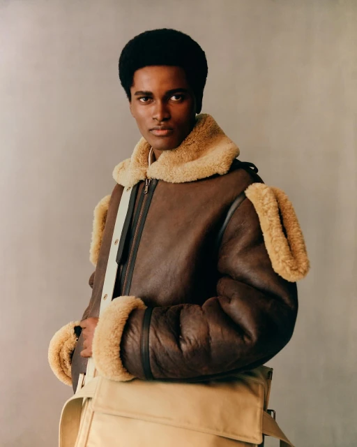 A stylish black male model poses indoors wearing a brown shearling bomber jacket and a beige messenger bag in a fashion editorial shot.