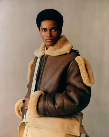 A stylish black male model poses indoors wearing a brown shearling bomber jacket and a beige messenger bag in a fashion editorial shot.