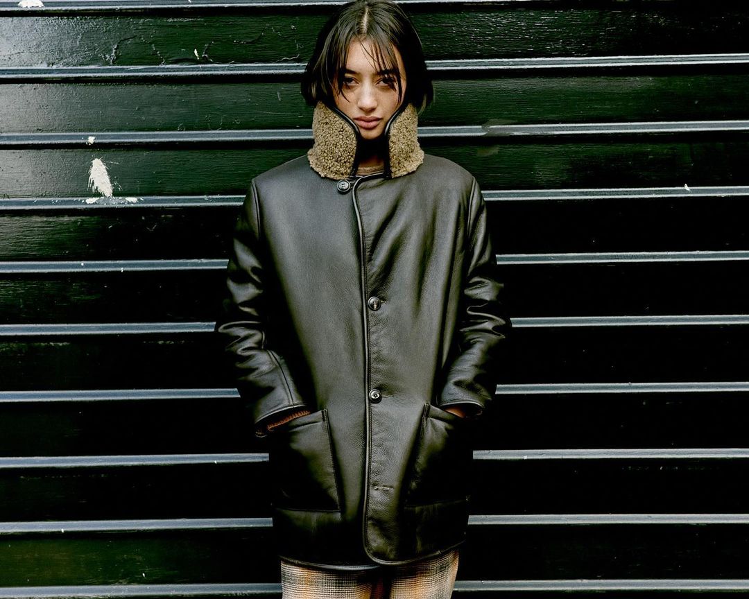 Woman in a dark leather jacket with a fur collar poses against a dark, horizontally-striped wall in a medium, eye-level shot.