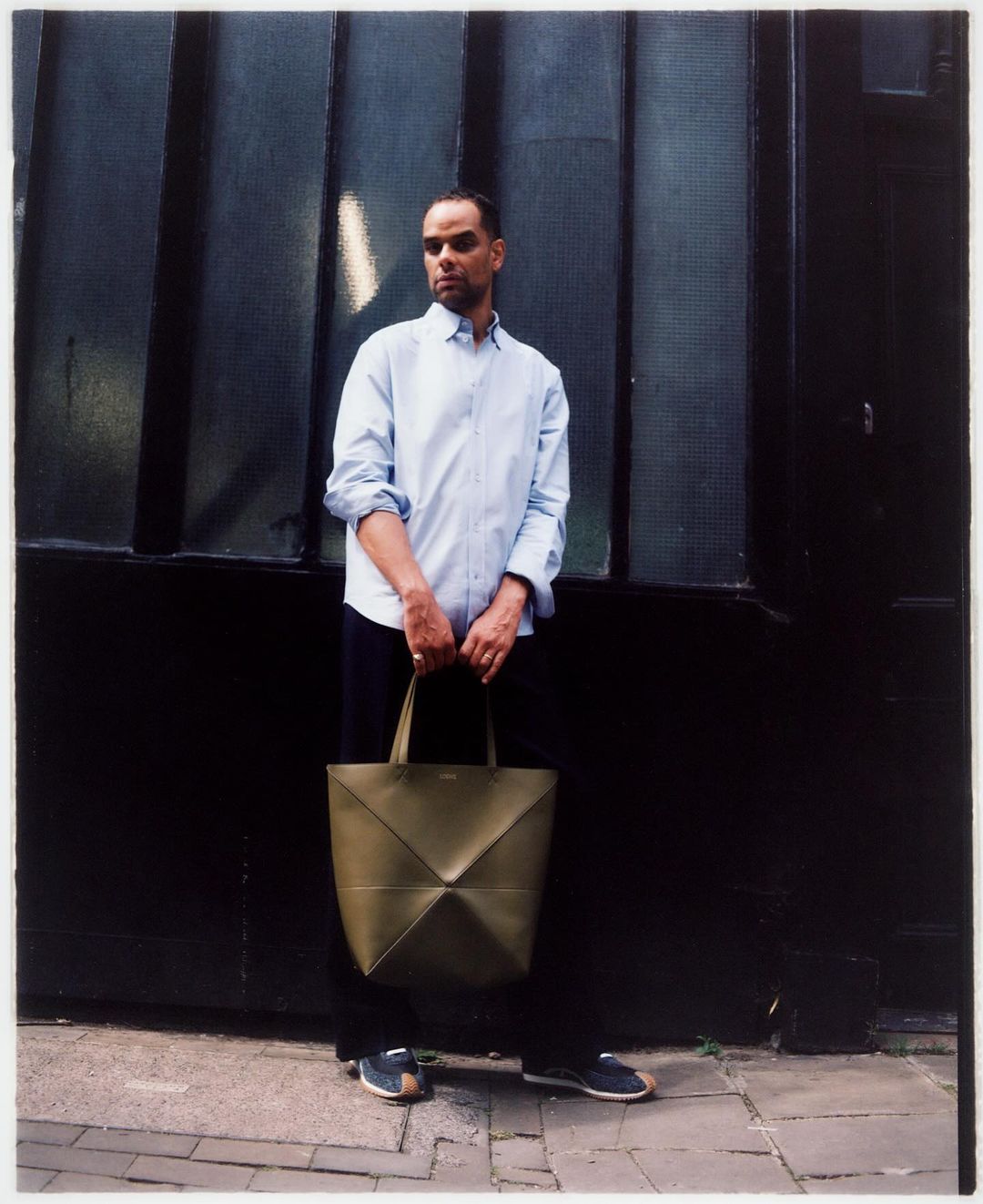 Man with olive green tote bag standing against a dark paneled wall in an outdoor setting on a sunny day, creating a lifestyle fashion image.