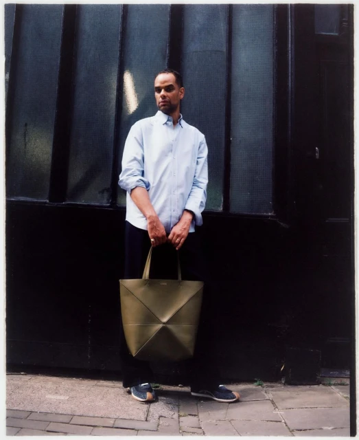 Man with olive green tote bag standing against a dark paneled wall in an outdoor setting on a sunny day, creating a lifestyle fashion image.