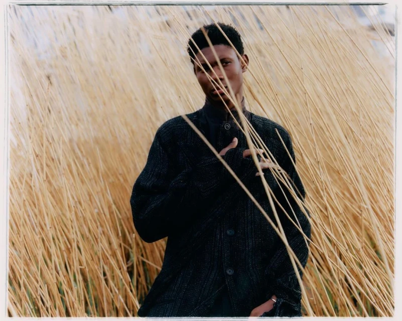 Portrait of an African American man in a dark jacket standing amongst tall, tan grasses in a natural outdoor setting.