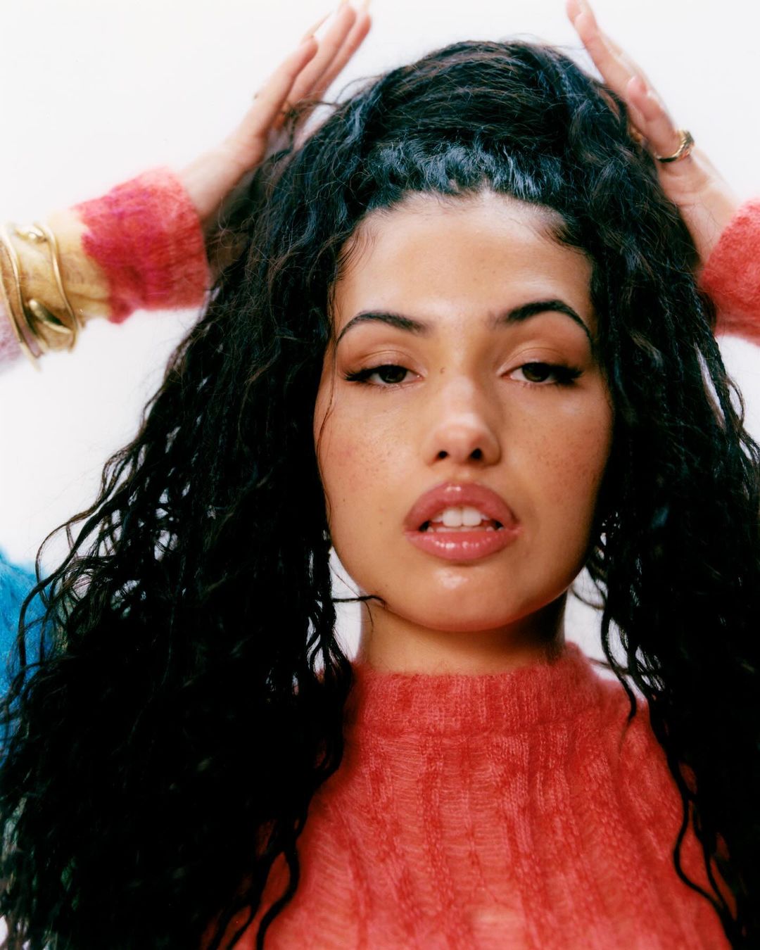 Close-up portrait of a woman with long dark curly hair wearing a pink textured sweater, photographed by Arthur Comely in London.