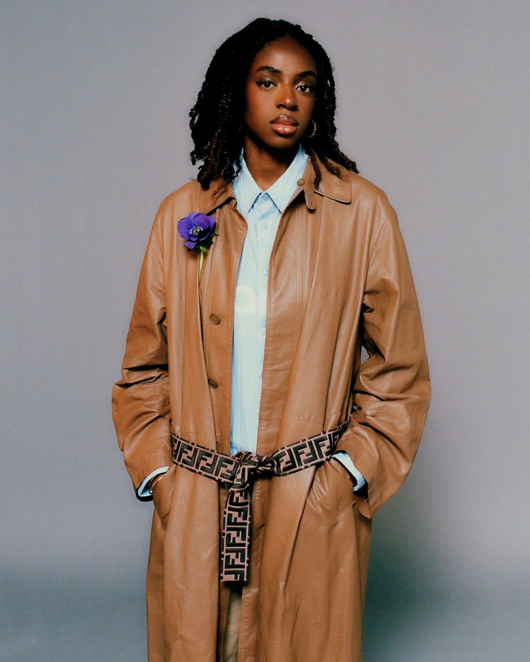 Full shot of a stylish black woman in a tan coat with a logo belt, light blue shirt, and purple flower posing in front of a grey background.