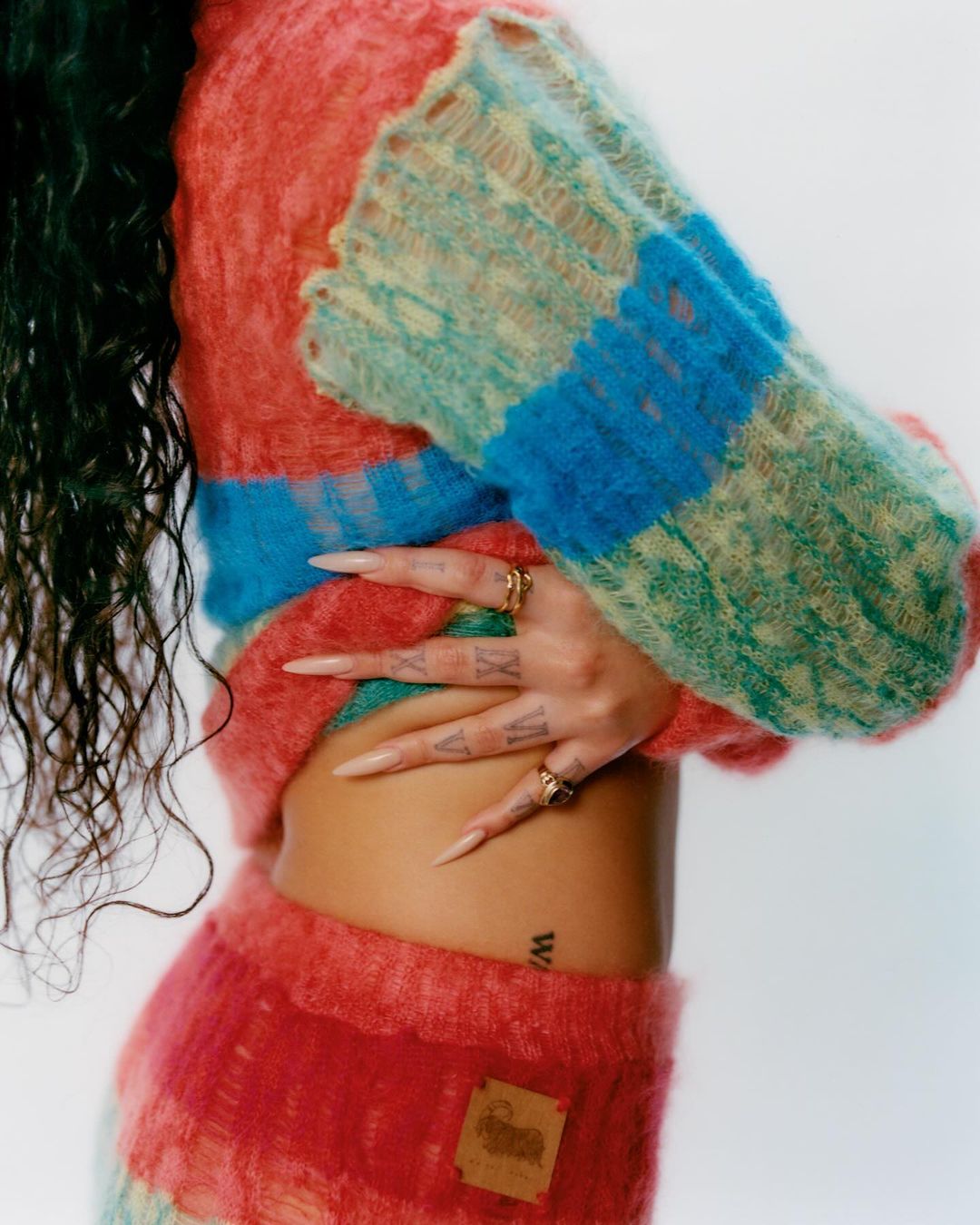 Woman wearing a colorful knitted sweater and skirt poses indoors. The photo features details of the soft fabric and her long fingernails.