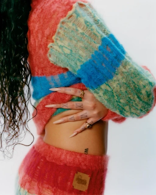 Woman wearing a colorful knitted sweater and skirt poses indoors. The photo features details of the soft fabric and her long fingernails.