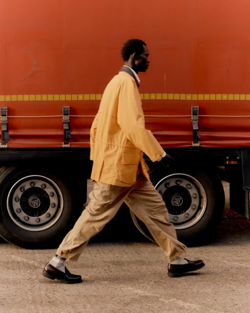 A dark-skinned man in a tan jacket and pants walks in profile in front of a large orange truck, showcasing fashion and urban style.