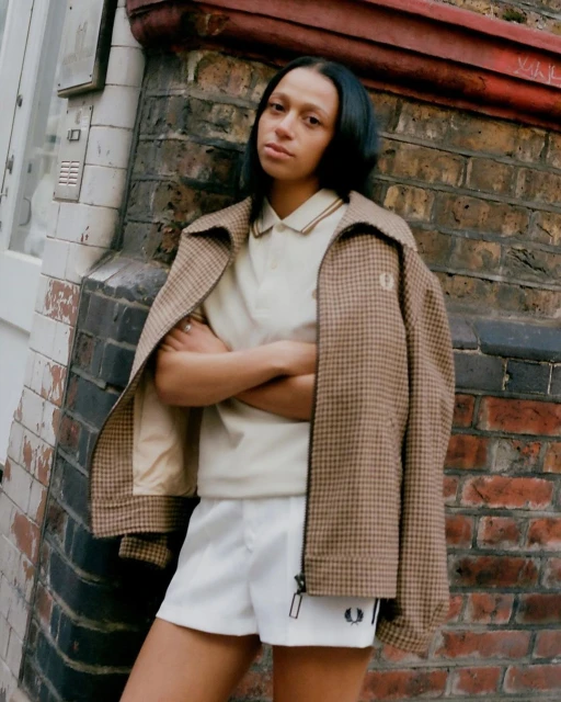 An adult woman poses outdoors, wearing a tan jacket, polo shirt, and white shorts, leaning against a brick wall with neutral expression.