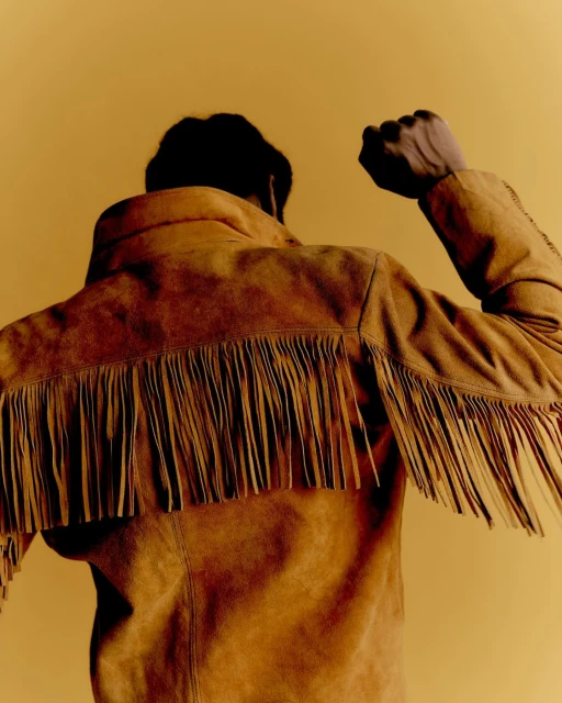 Back view of a person wearing a fringed tan suede jacket, arm raised in a fist, against a warm yellow-toned background in a studio setting.