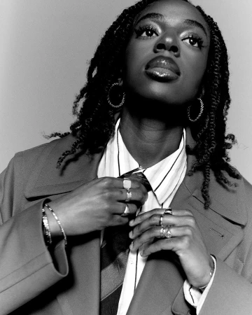 Black woman adjusting her tie wearing a striped shirt and blazer with braided hair and hoop earrings in a monochrome portrait.