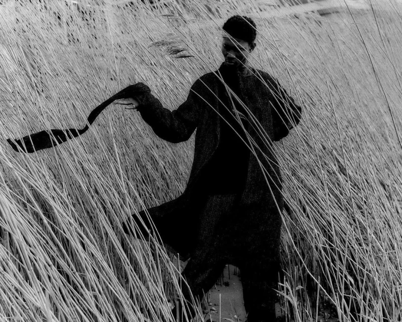 Black and white portrait of an African male in a coat, holding a scarf, and standing in a field of tall grass.