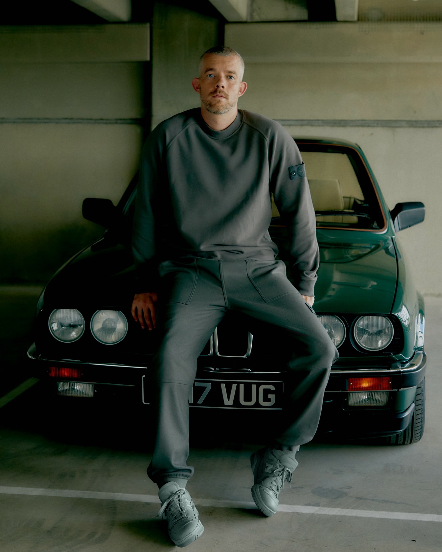 Man in grey sweatsuit seated on green BMW in garage with concrete ceiling and floor poses for fashion photography.