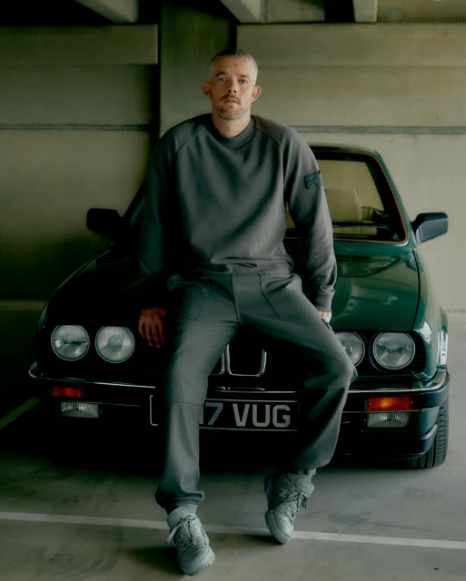 Man in grey sweatsuit seated on green BMW in garage with concrete ceiling and floor poses for fashion photography.