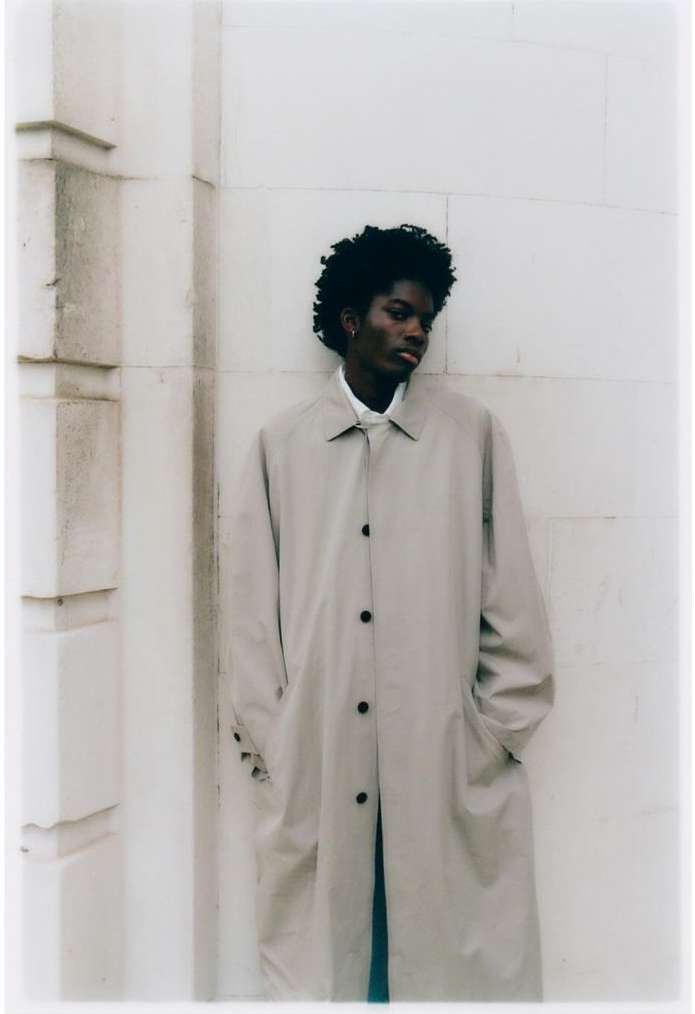 A slender Black male stands against a textured wall in a tan trench coat, his gaze directed off-frame, creating a composed and stylish look.