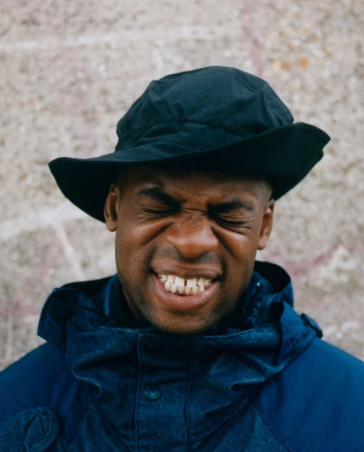 Close-up of a Black adult male wearing a dark hat and blue jacket, squinting with a closed-mouth smile against a textured wall backdrop.