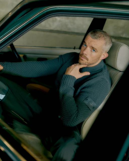 Adult male poses in a car wearing a textured sweater with a brand patch on the sleeve; interior shot with natural light.