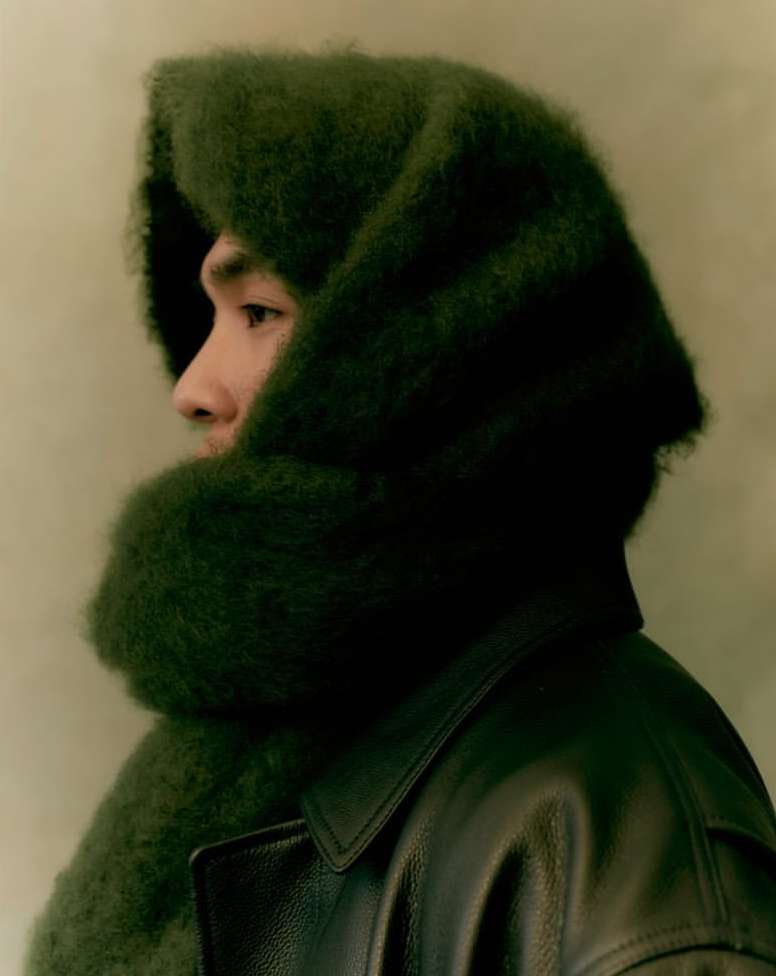 Portrait of an Asian man wearing a fuzzy, green hooded scarf and dark jacket, looking off to the side with a neutral expression.