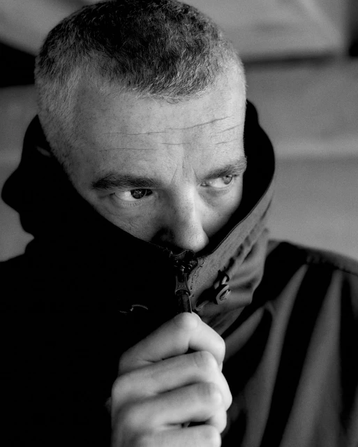 Black and white portrait of a man with short grey hair, partially obscured by a dark hooded jacket, looking to the side with a serious expression.
