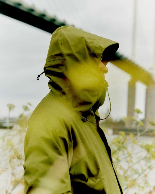 Profile shot of a person wearing a green hooded raincoat, standing outside near a bridge on an overcast day. Focus on the figure and coat.