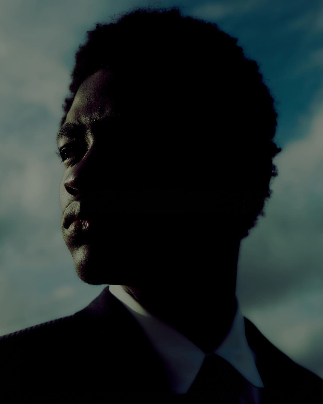 Dramatic close-up portrait of an African American man in a suit against a cloudy sky, defined by dark shadows and cool tones.