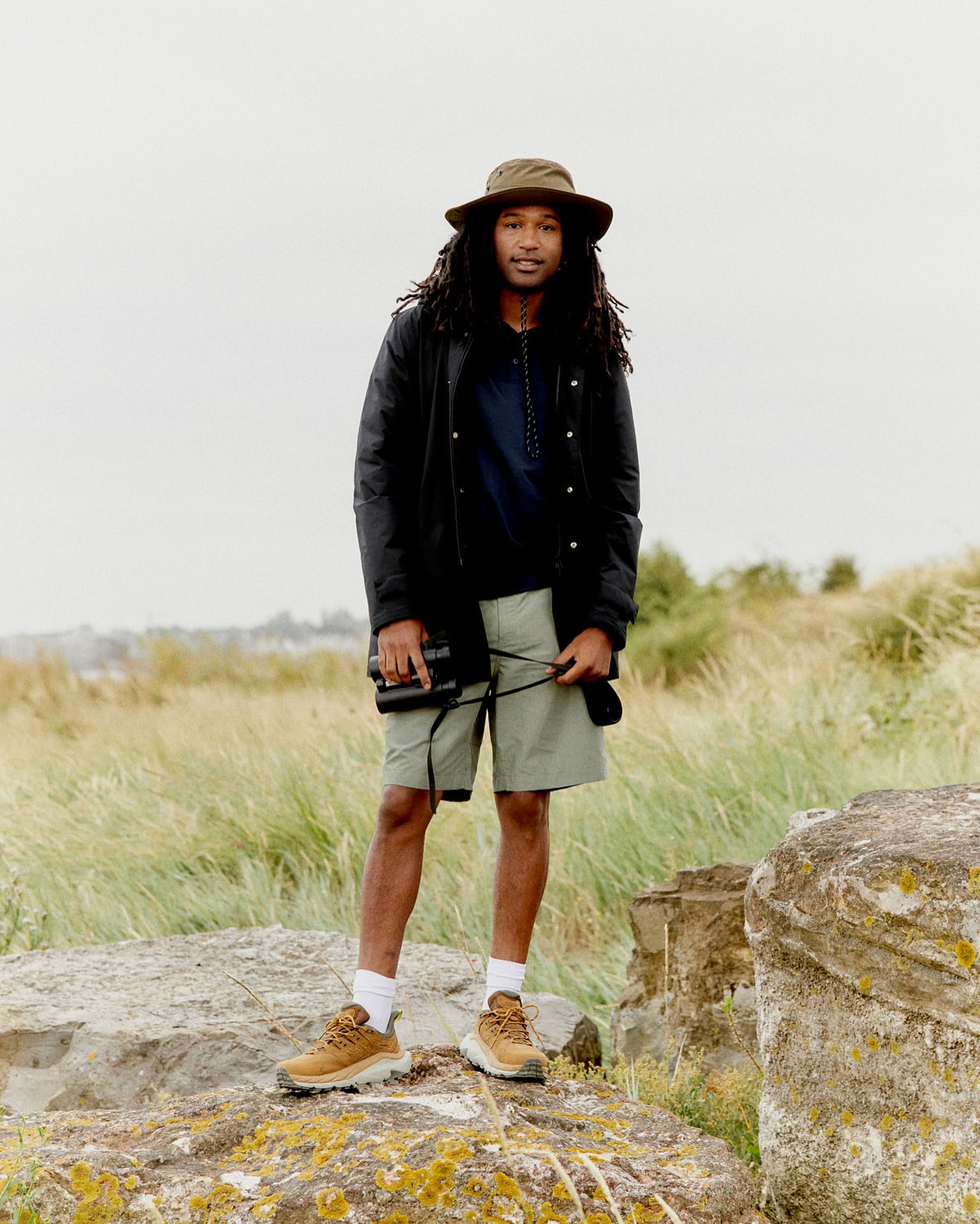 A man standing on rocks in outdoor apparel. He's wearing a hat, jacket, and shorts, holding binoculars, with grass and cloudy sky behind him.