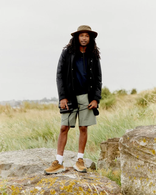 A man standing on rocks in outdoor apparel. He's wearing a hat, jacket, and shorts, holding binoculars, with grass and cloudy sky behind him.