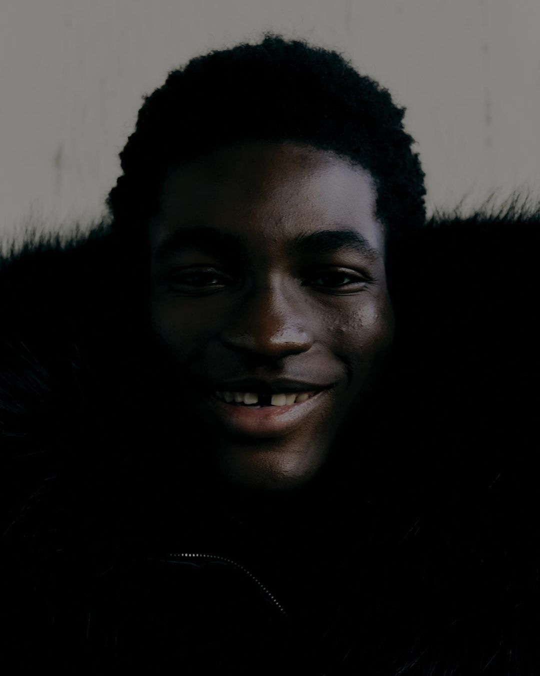 Portrait of a smiling, young, Black man with dark curly hair, wearing a fur-trimmed jacket, against a neutral background.