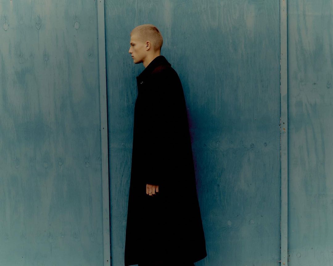 Profile shot of a short-haired, fair-skinned person in a long black coat standing against a textured blue wall indoors.
