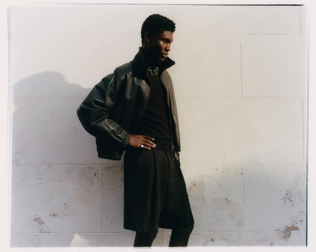 A dark-skinned male model poses with his hand on his hip, wearing a leather jacket, against a white textured wall in a fashion editorial shoot.