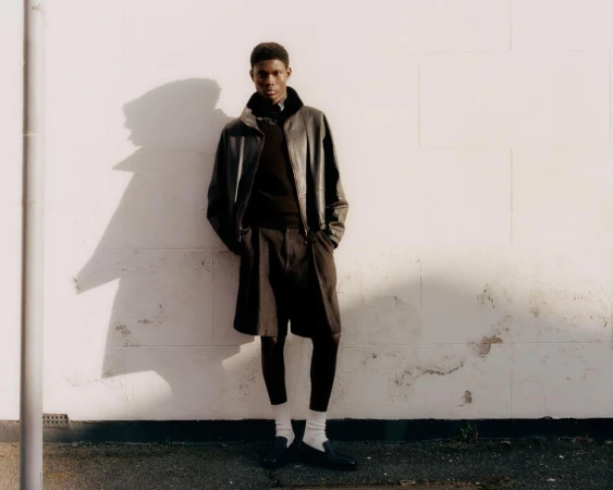 Young Black man wearing a black leather jacket and dark shorts leaning against a light exterior wall outdoors.