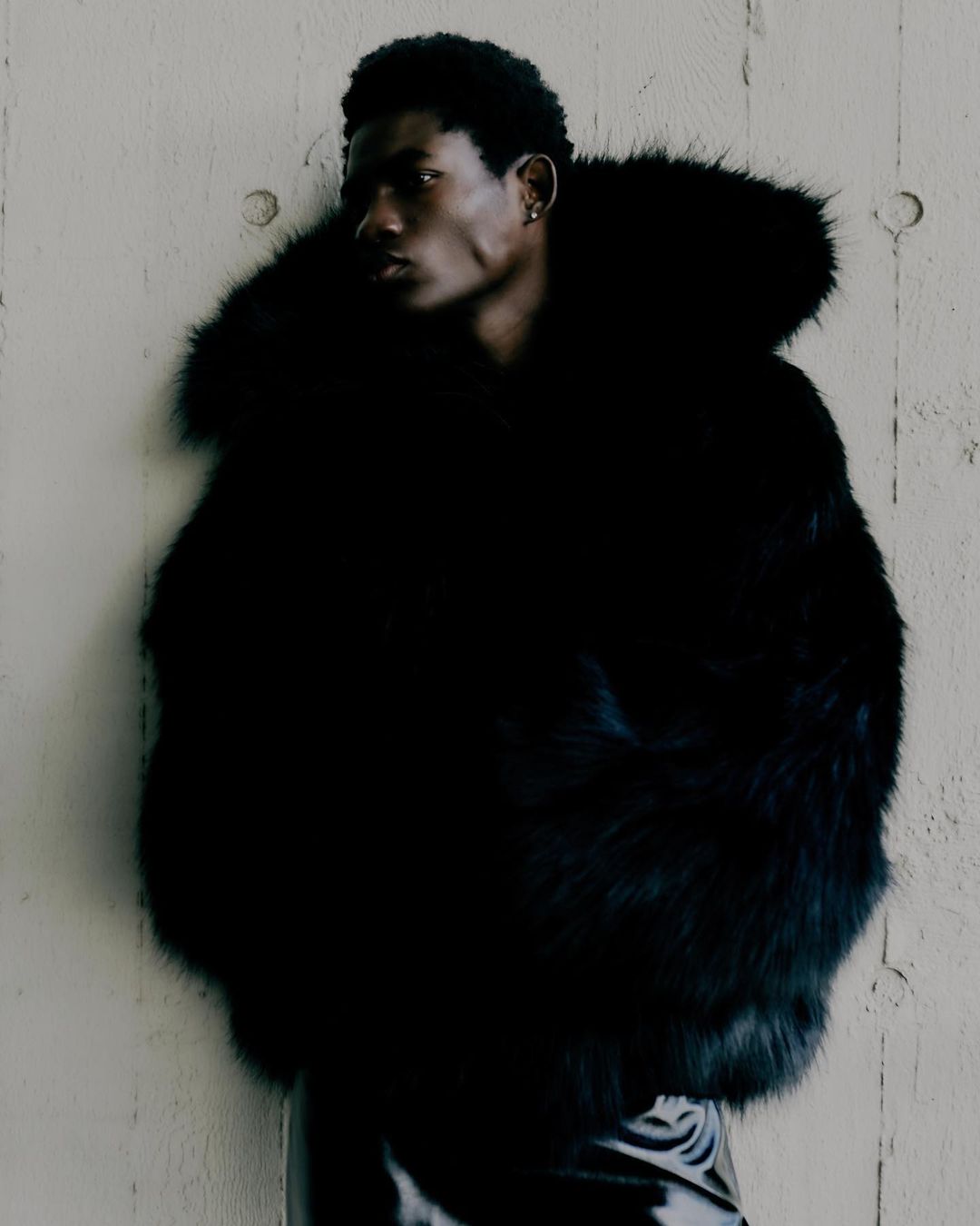 Portrait of a dark-skinned man wearing a large, black fur coat, posing against a textured wall. Fashion editorial style.