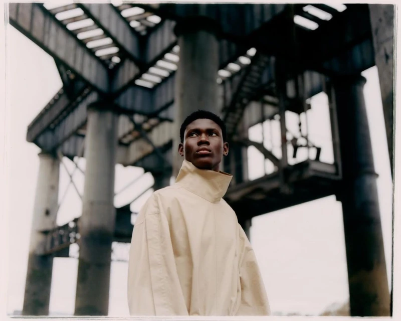 Young man with dark skin poses in a beige top with a high collar, standing under a concrete industrial structure on an overcast day.