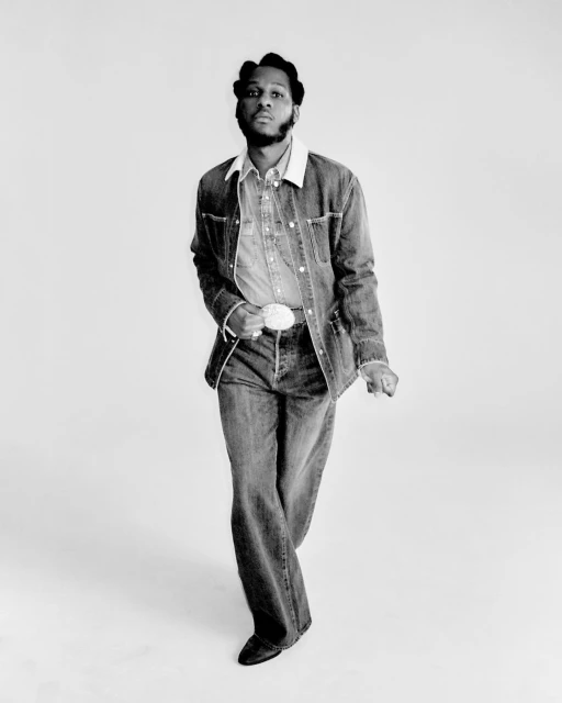 A stylish Black male in denim jacket and bell-bottom jeans poses in a full shot against a white background in a black-and-white studio portrait.