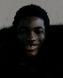 Close-up portrait of a young Black man smiling, wearing a dark faux fur jacket against a pale wall.
