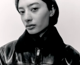 Close-up black and white portrait of a model wearing a dark leather jacket with a high collar, photographed by Arthur Comely.