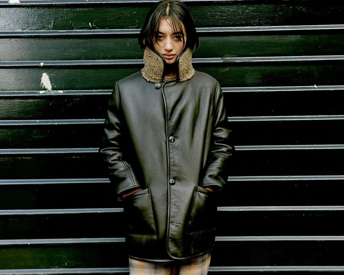 A person wearing a dark leather coat with a shearling collar standing against a dark slatted background.