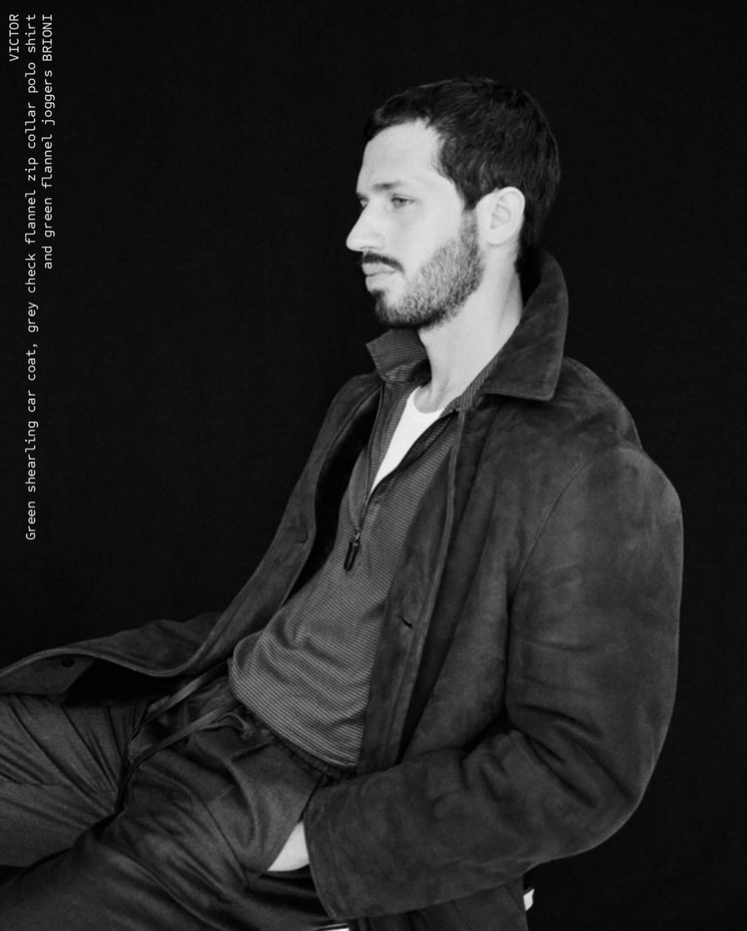 A man with a beard poses in a car coat, flannel shirt, and joggers against a dark background. He has a hand in his pocket.